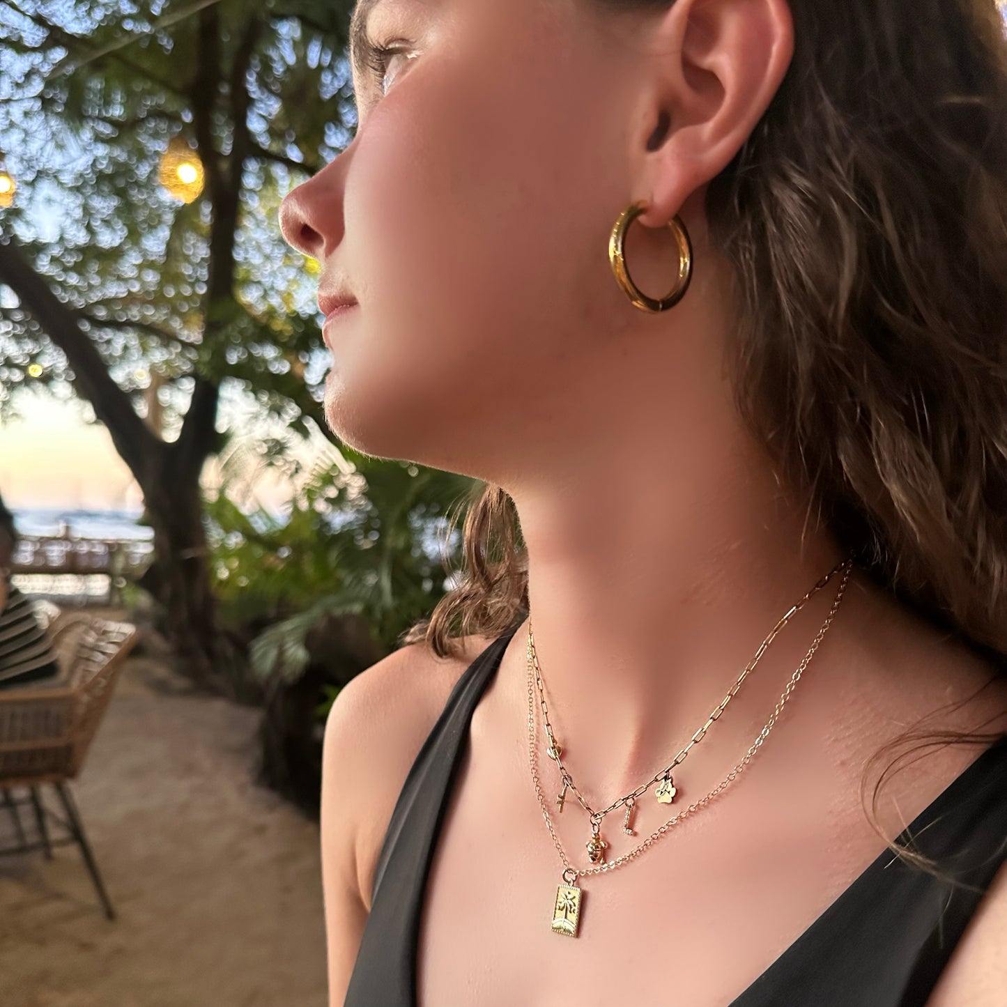 Woman wearing gold hoop earrings and necklaces with a blurred beach background