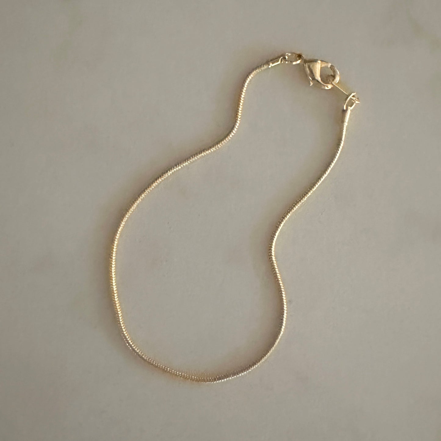 A delicate gold chain necklace with a lobster clasp, laid out on a light beige marble surface.