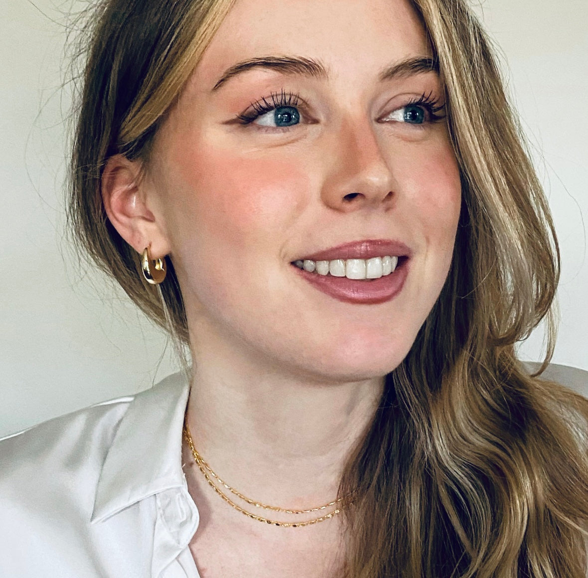 A close-up portrait of a smiling young woman with blonde hair, wearing a small gold hoop earring and delicate gold chain necklaces, against a plain light background.