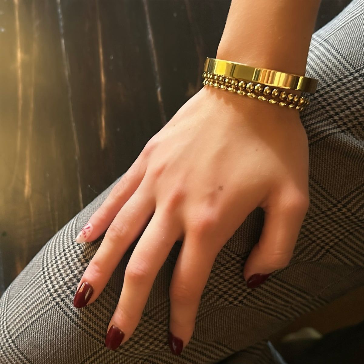 A close-up of a person's hand resting on gray plaid fabric, wearing a wide gold cuff bracelet with two rows of small metallic beads.