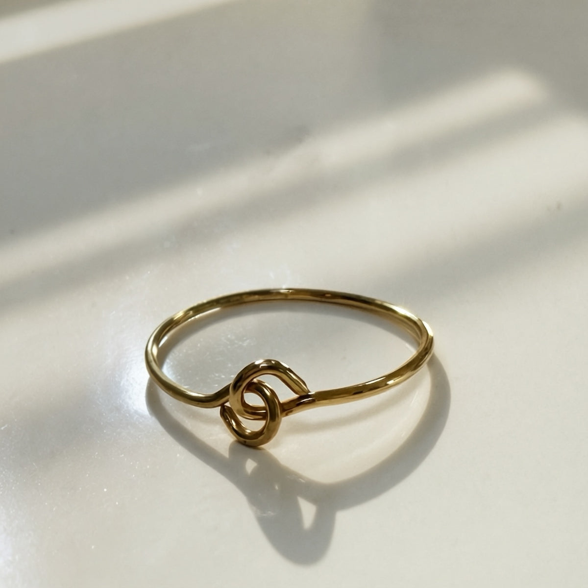 A delicate gold wire ring with a stylized knot design, resting on a white surface with soft window shadows.