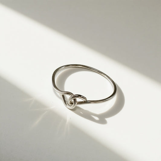 A minimalist sterling silver ring with a unique interlocking knot design, photographed on a white surface under dramatic diagonal sunlight and shadow.