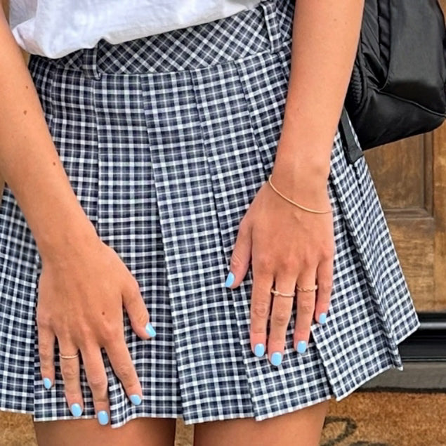 A close-up of a pleated mini skirt with a navy and white gingham plaid pattern, worn by a model with light blue nail polish and a gold bracelet.
