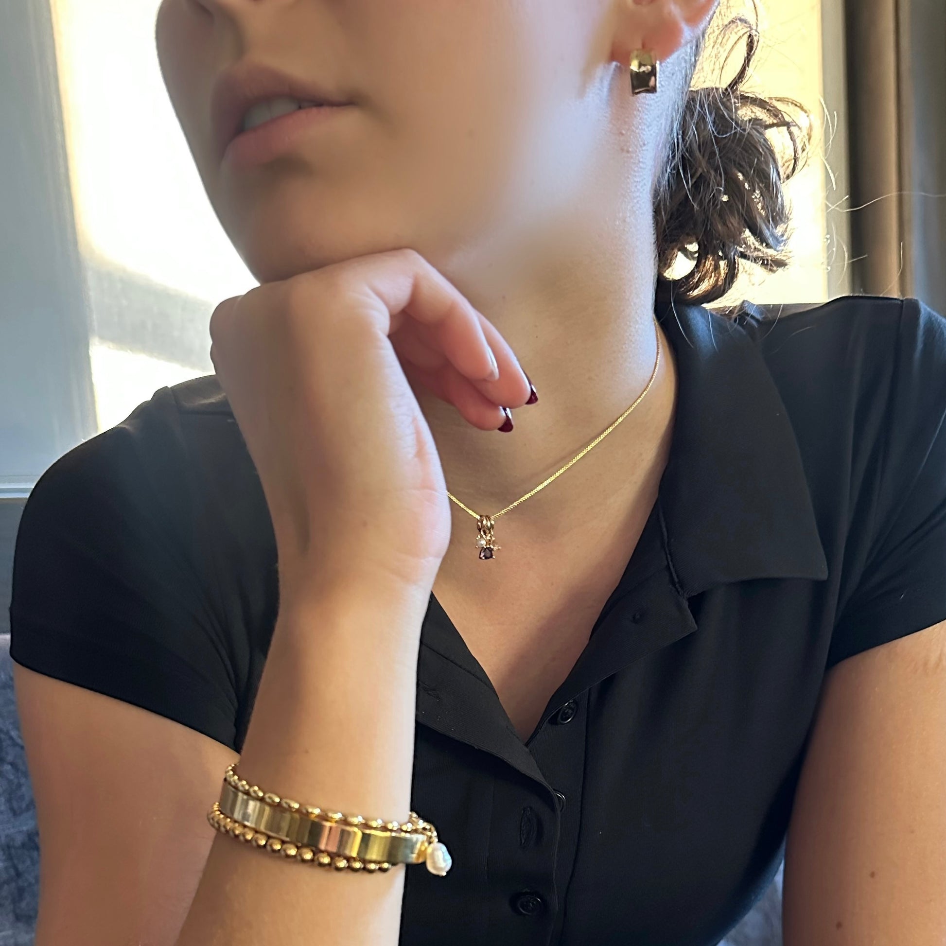 A close-up lifestyle shot of a person wearing a delicate gold chain necklace with a small dark pendant, a gold beaded bracelet, and a black collared shirt.