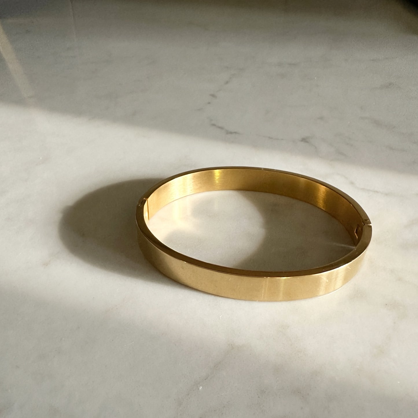 A polished gold-colored metal bangle bracelet resting on a white marble surface with soft natural shadows.