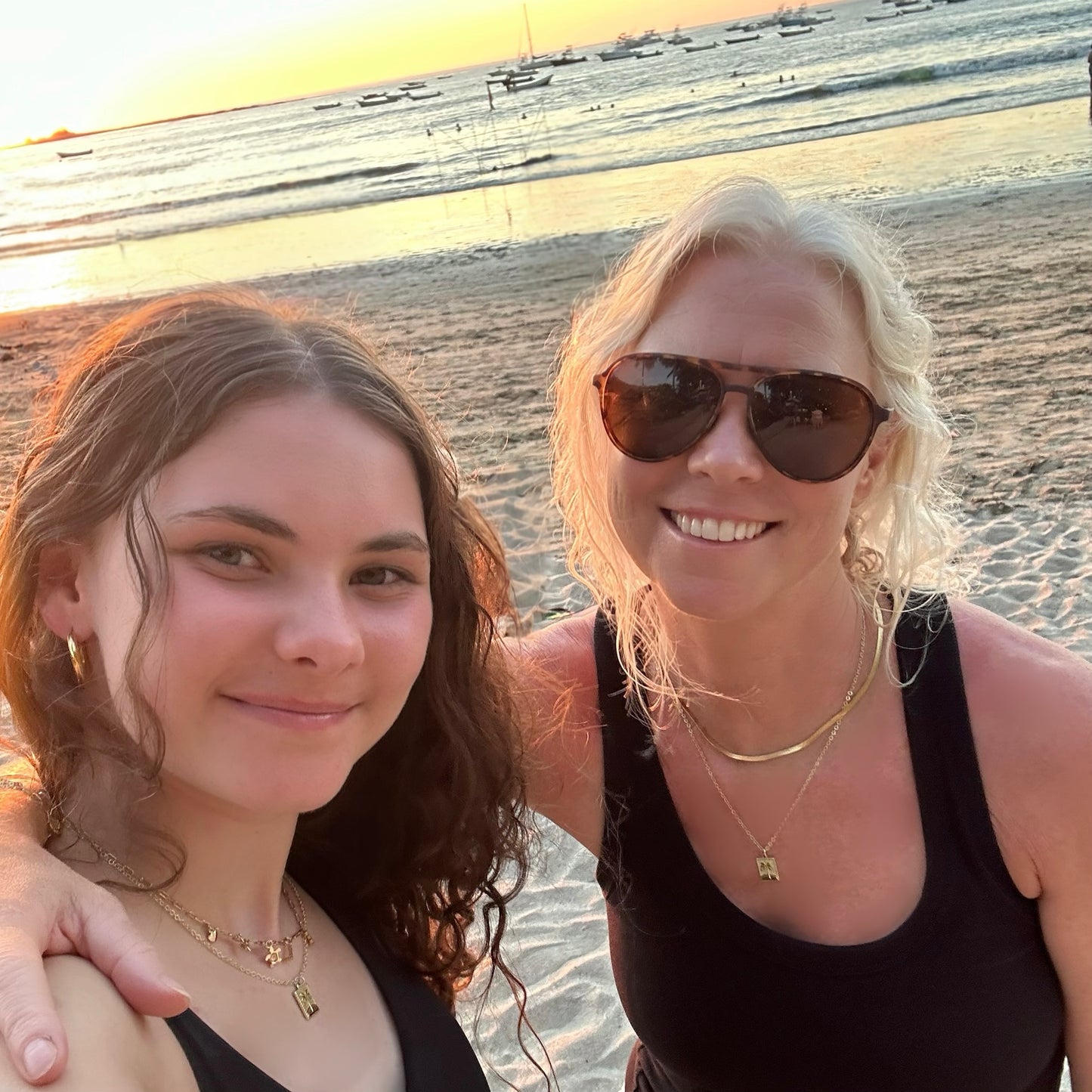 Two women posing for a photo on a beach at sunset, each wearing a rectangle palm tree pedant