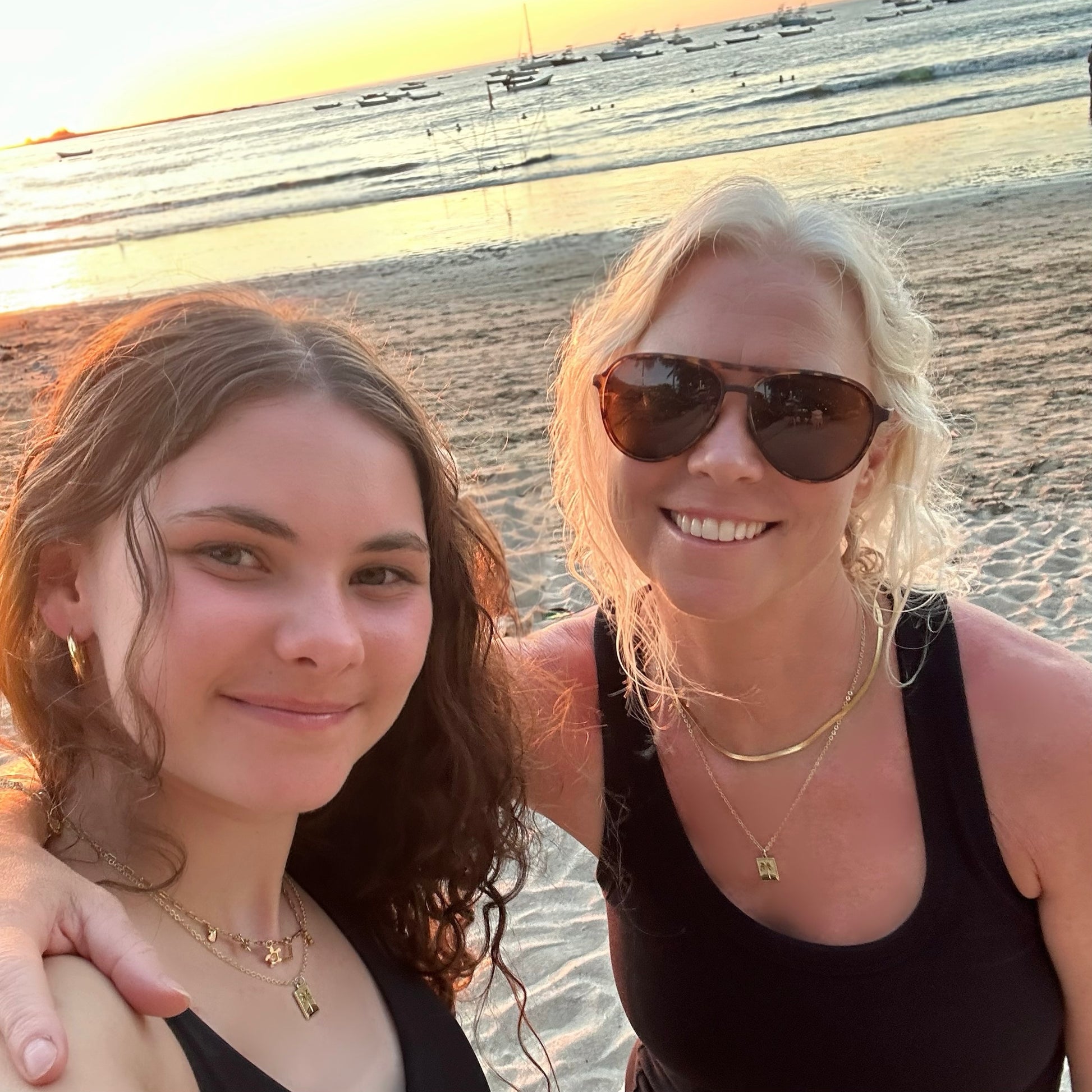 Two women posing for a photo on a beach at sunset, each wearing a rectangle palm tree pedant