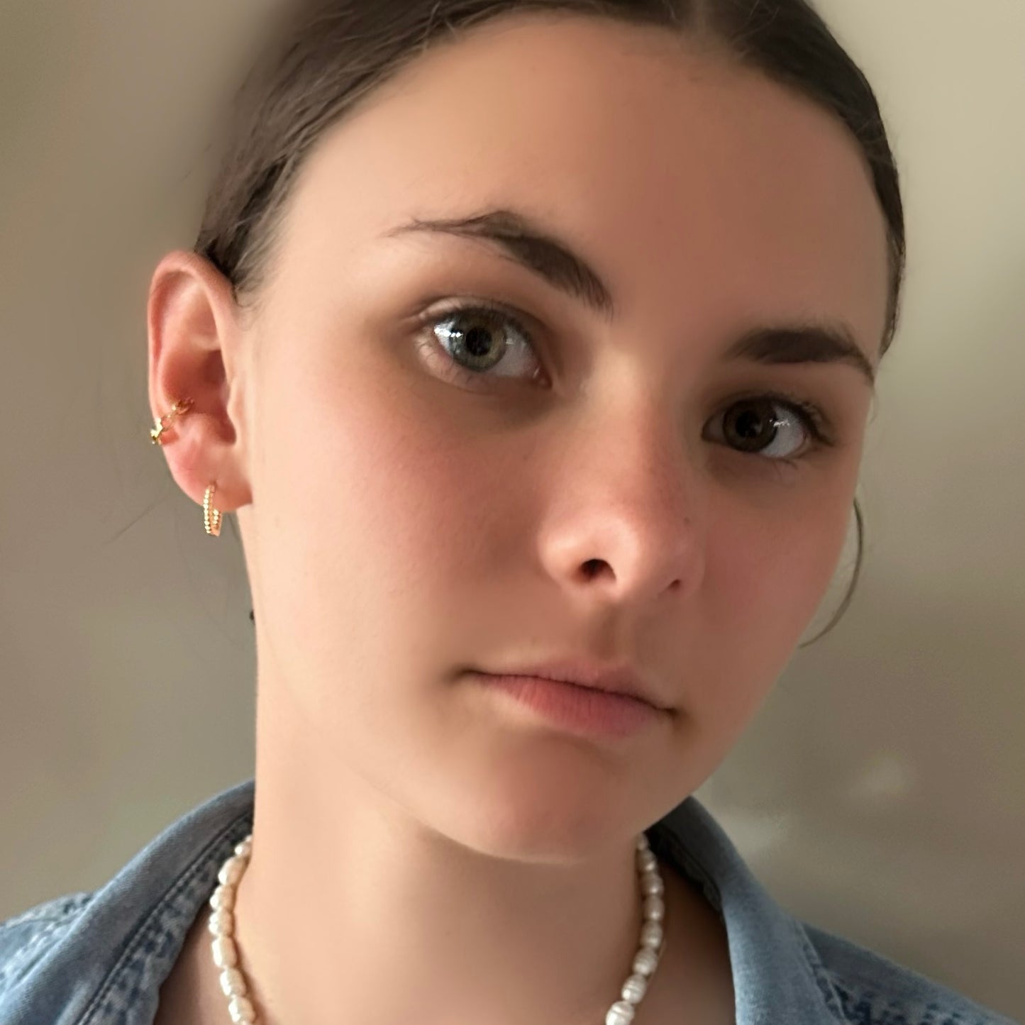 Close-up of a woman wearing gold hoop earrings and a pearl necklace against a neutral background