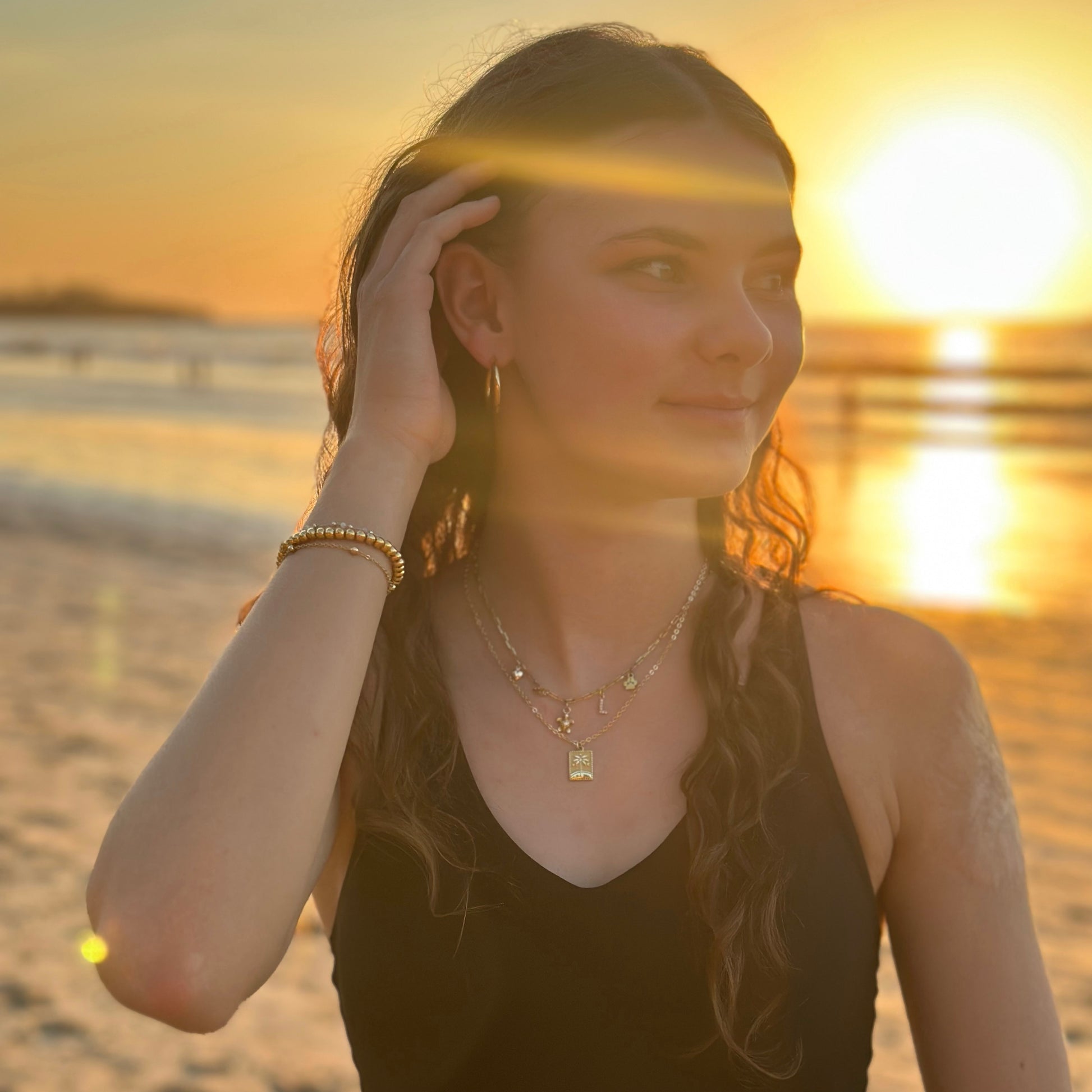 Woman on beach wearing a necklace with a rectangle palm tree pendant with the sunset in the background