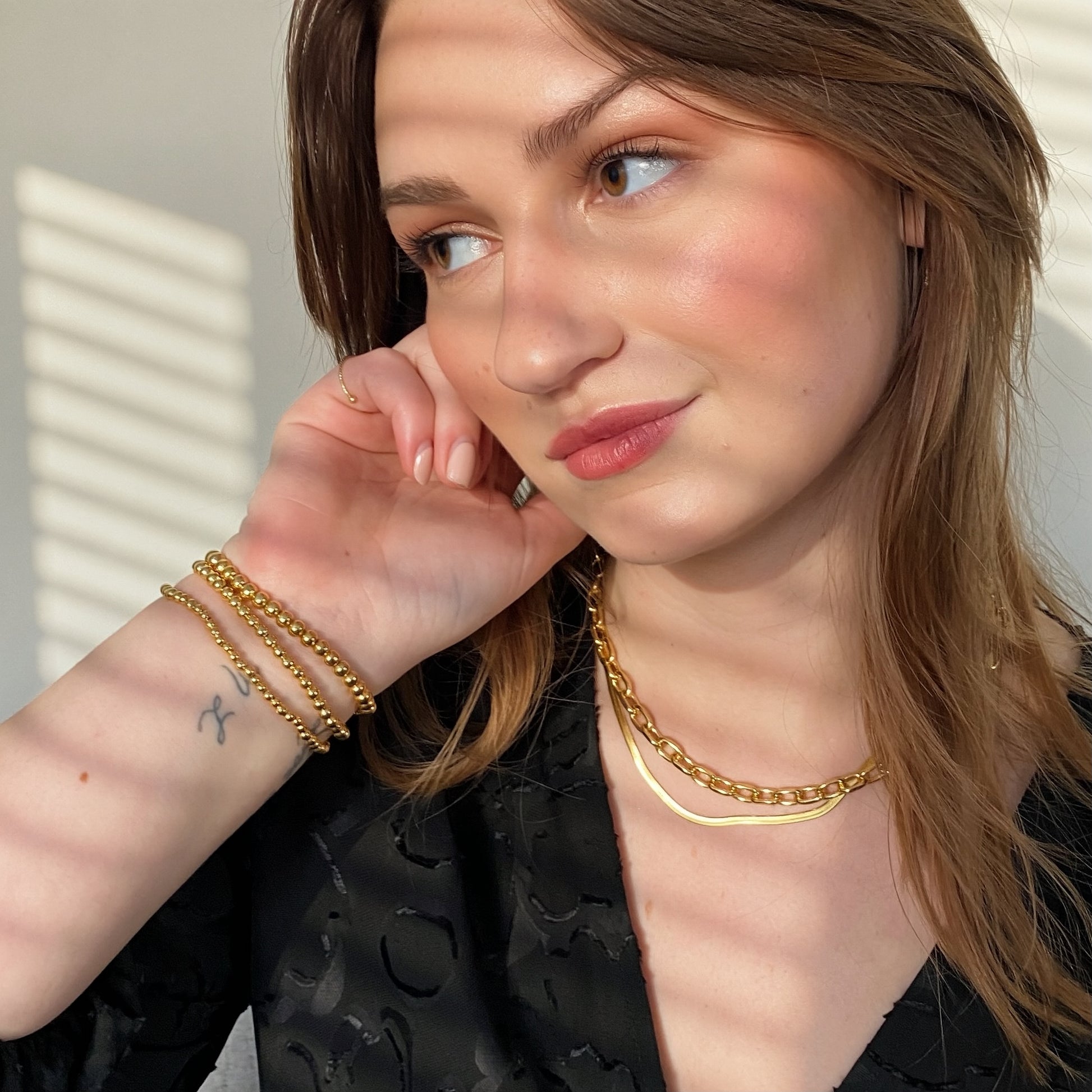 A close-up lifestyle shot of a woman wearing gold jewelry, including a layered stack of gold chain bracelets on one wrist and a gold chain necklace, paired with a black textured top.