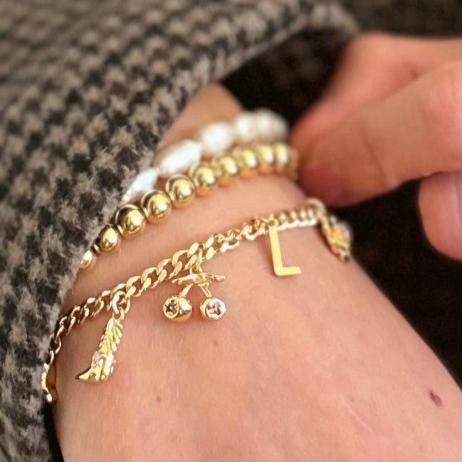 A close-up of a person's wrist wearing multiple gold-tone charm bracelets, including a chain bracelet with a feather, cherries, and letter L charm, layered over white pearl bead bracelets.