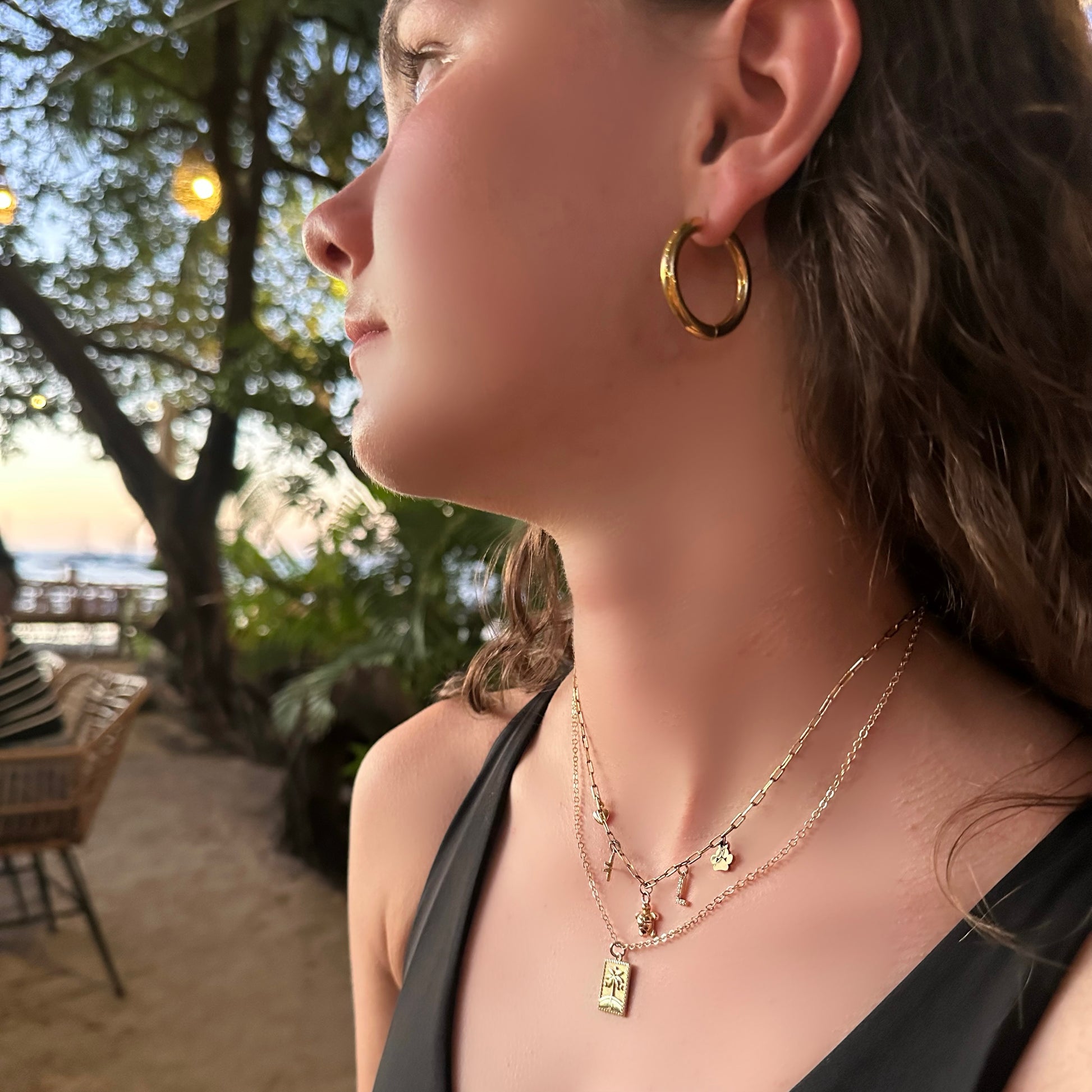 Woman wearing gold hoop earrings and necklaces with a blurred beach background