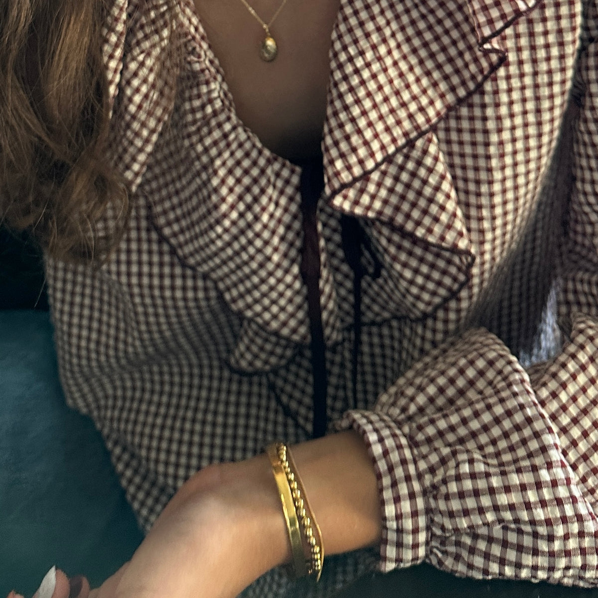 A close-up of a person wearing a brown and white gingham blouse with ruffled details and a black tie at the neckline, accessorized with gold bangles and a necklace.