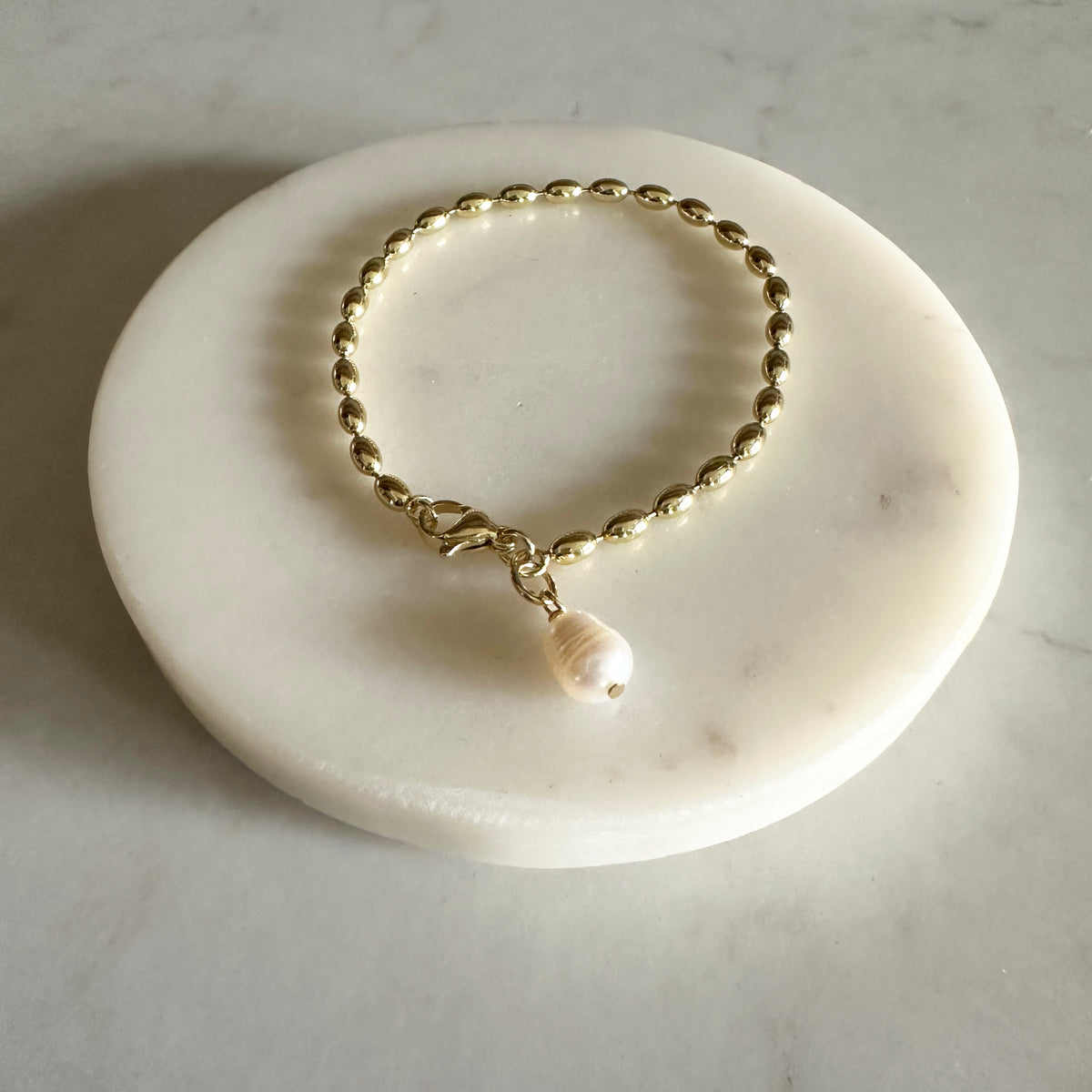 A gold-toned beaded chain bracelet with a small white pearl pendant, displayed on a round white marble stand against a light marble surface.