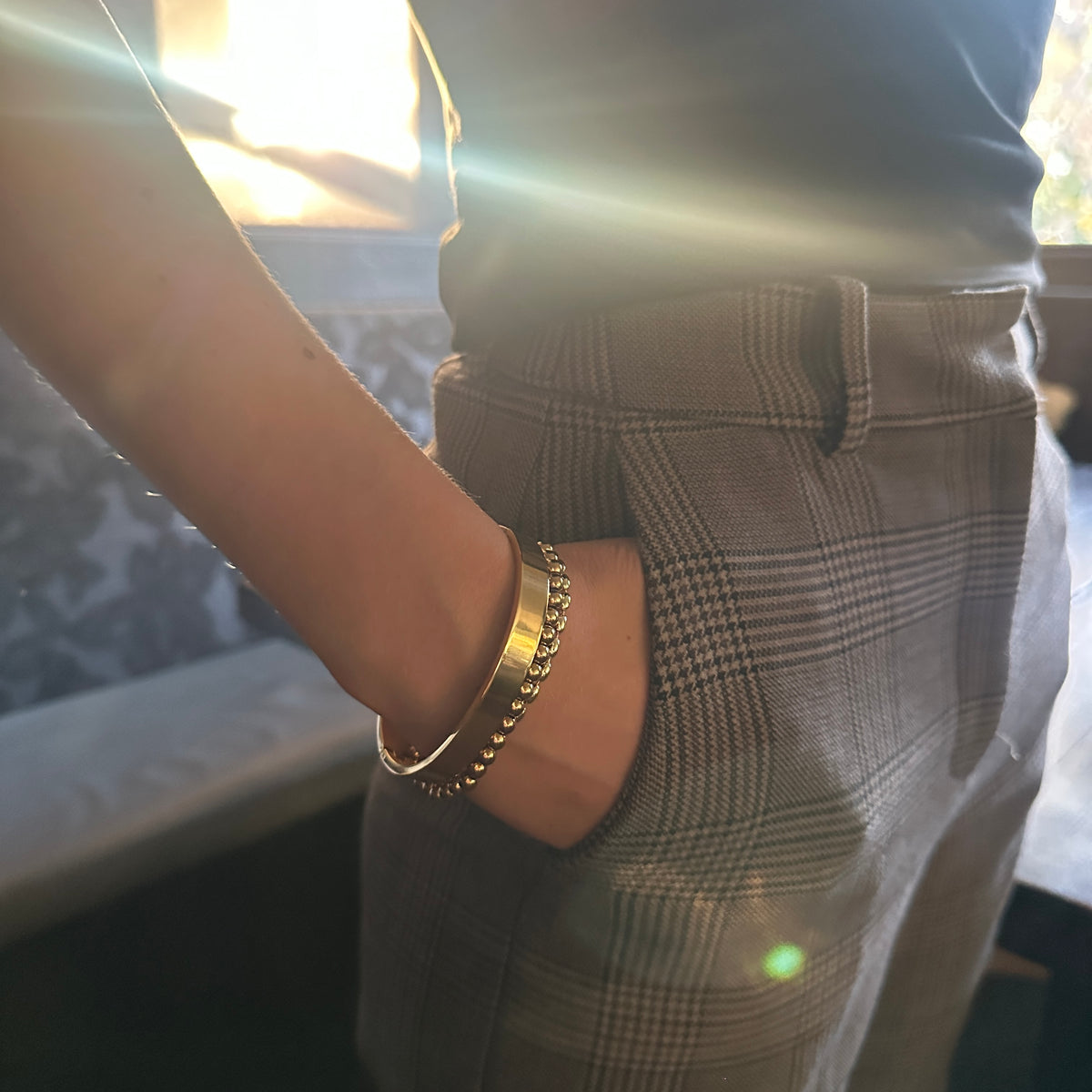 A close-up lifestyle shot of a person wearing a gold beaded bangle bracelet on their wrist, paired with brown plaid trousers and a dark top, under warm sunlight with lens flare.