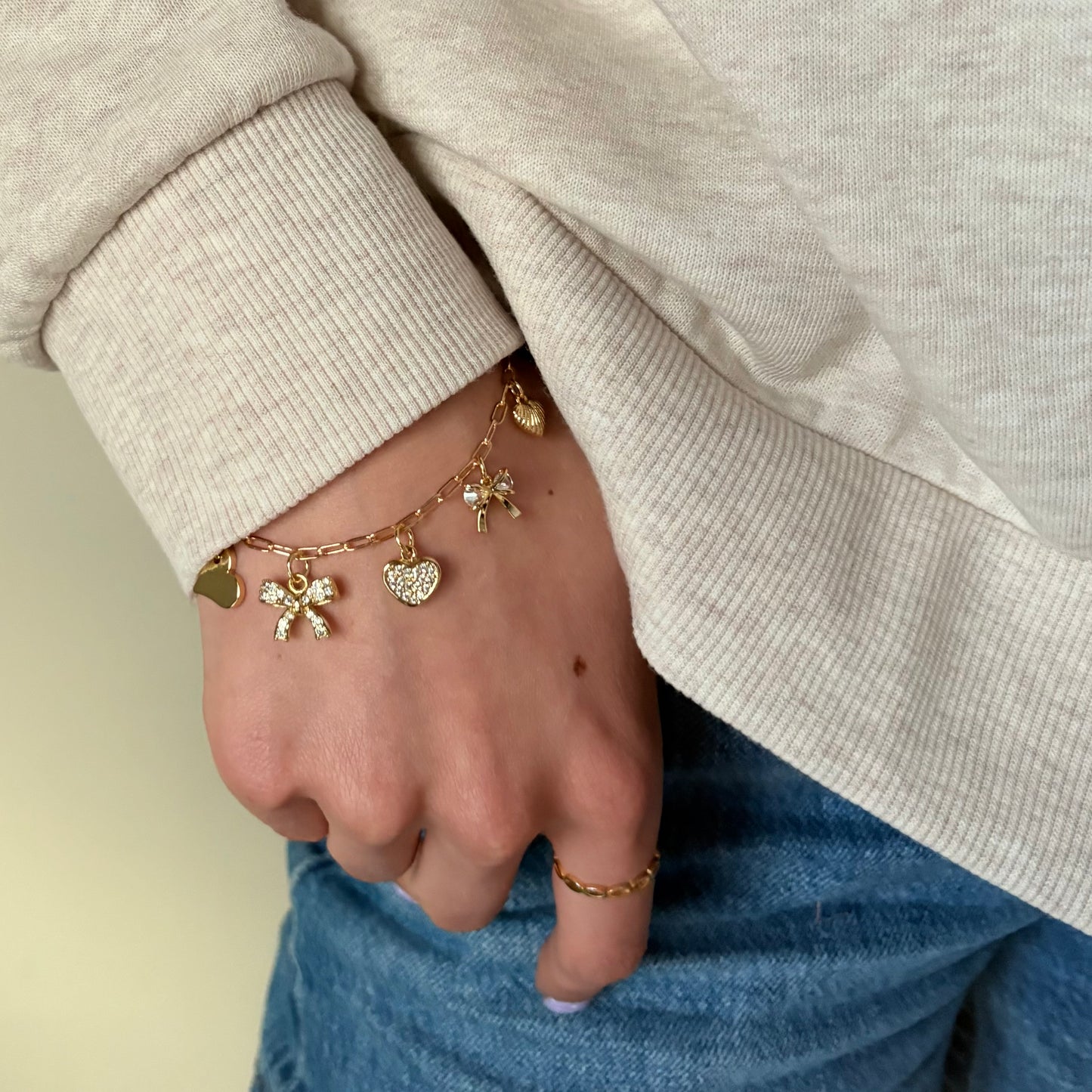 Hand wearing a gold bracelet with charms, wearing a beige sweater and blue jeans.