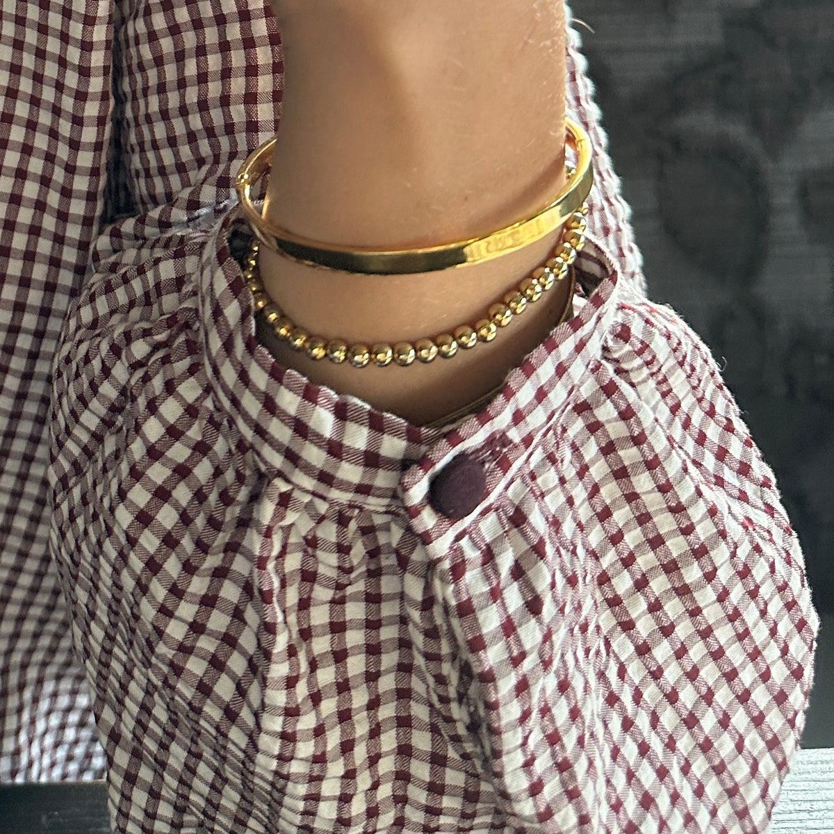 A close-up of a person's neck and wrist wearing three gold-toned bracelets: a thick smooth bangle, a thin chain bracelet, and a beaded chain bracelet, styled with a red and white gingham blouse.