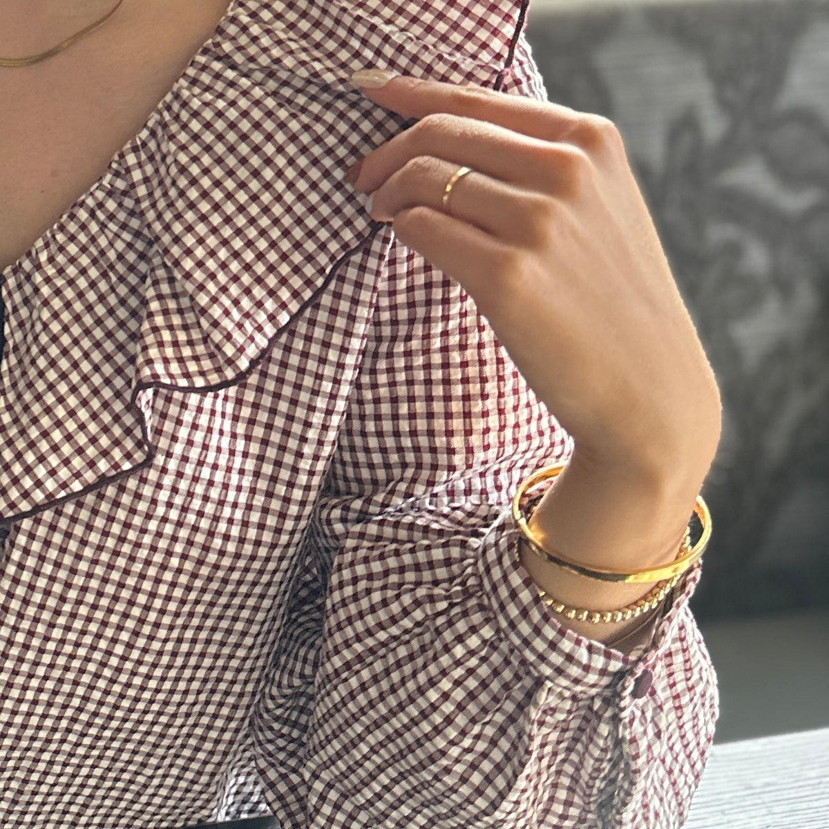 A close-up of a person wearing a red and white gingham check blouse, with a gold ring on their finger and stacked gold bracelets on their wrist.
