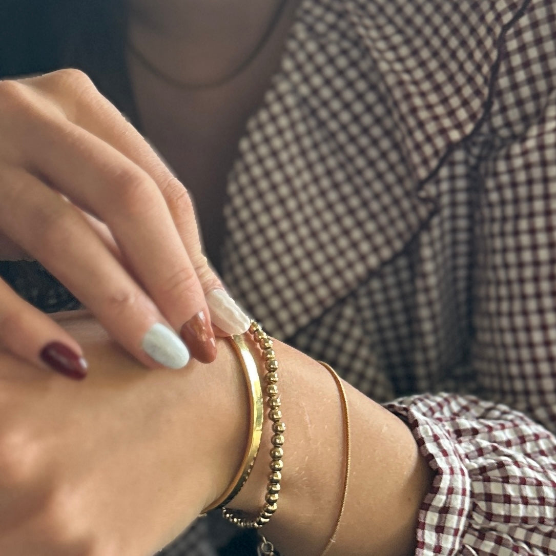 A close-up of a person's wrist wearing multiple delicate bracelets, including a chunky gold bangle, a beaded gold bracelet, and a thin gold chain, paired with a brown and white gingham shirt.