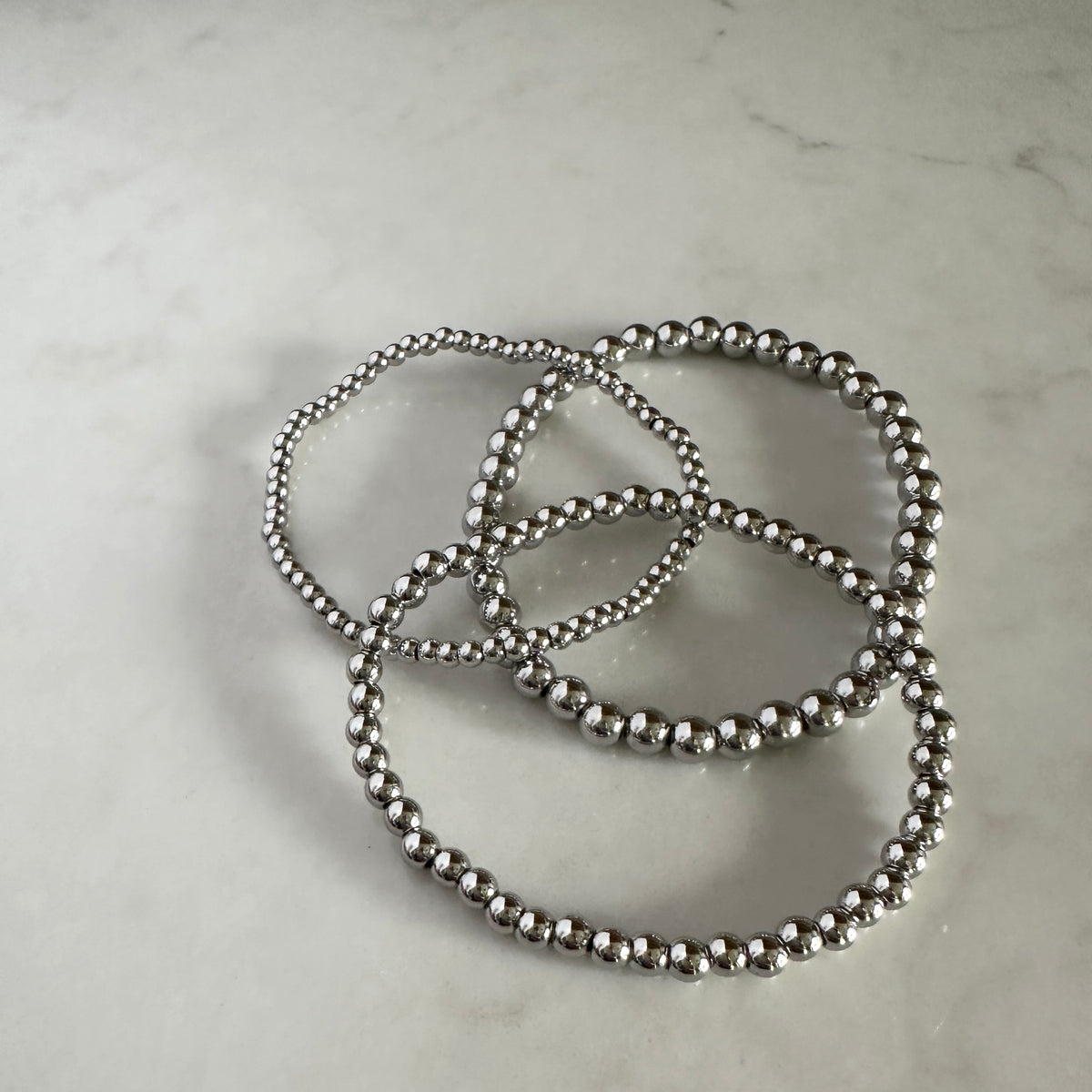 Three metallic silver beaded bracelets arranged in loops on a light gray marble surface.