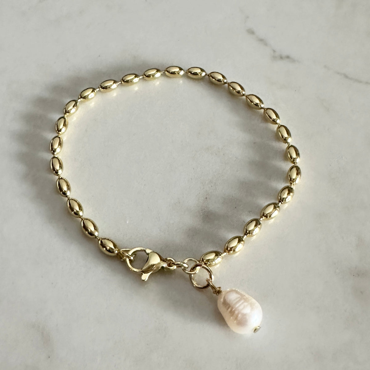 A gold chain bracelet with a small white pearl pendant, displayed on a light gray marble surface.