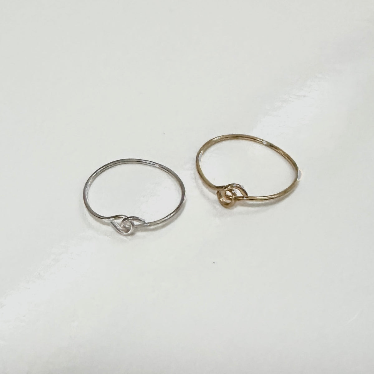 Two minimalist wire rings with knot-like details, one in silver and one in gold, displayed on a plain white surface.