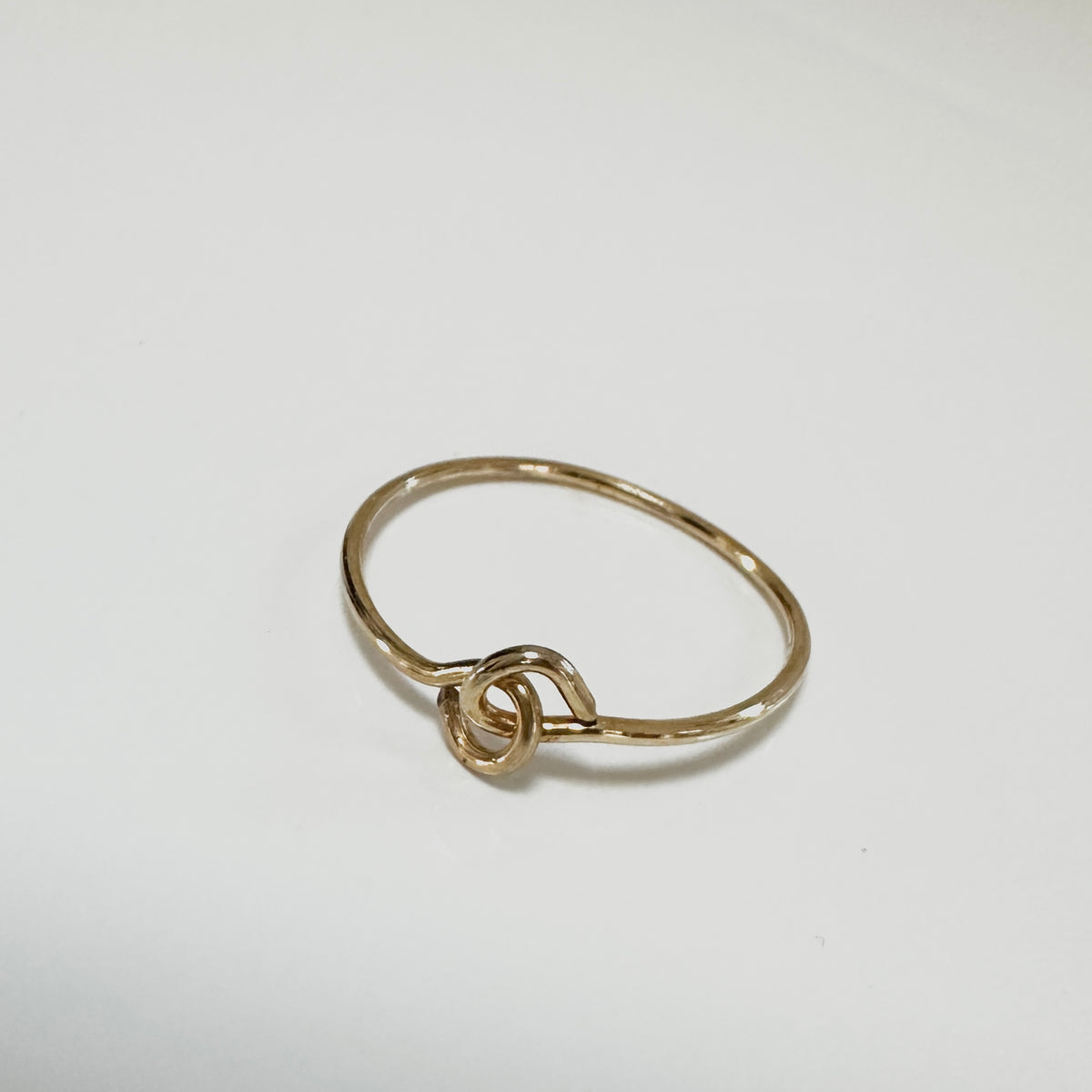 A thin gold ring with a knotted or interlaced loop detail at the front, photographed on a plain light background.
