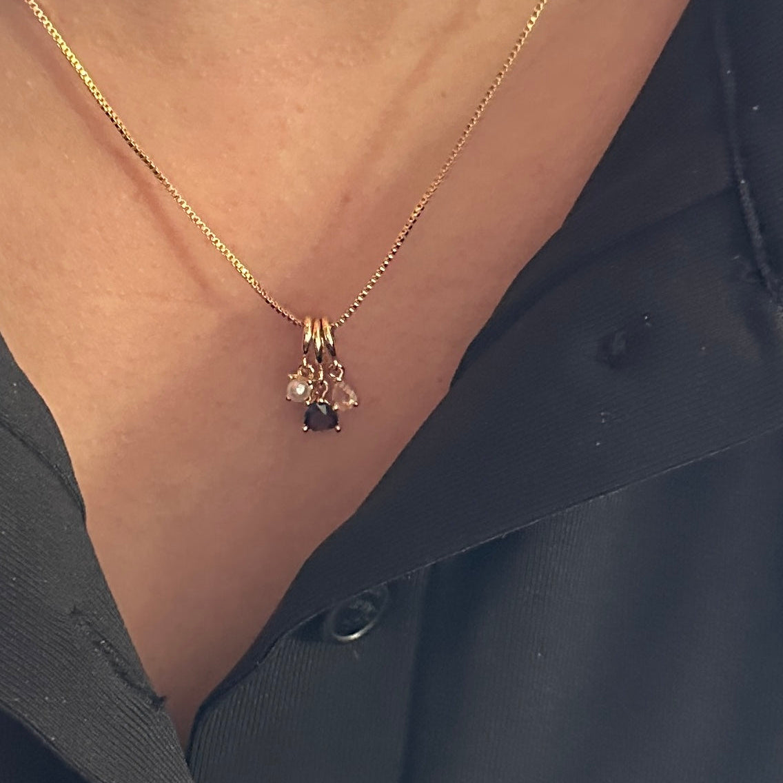 A close-up of a person wearing a delicate gold chain necklace with a small cluster pendant featuring a dark oval stone and clear stones, styled with a black collared shirt.