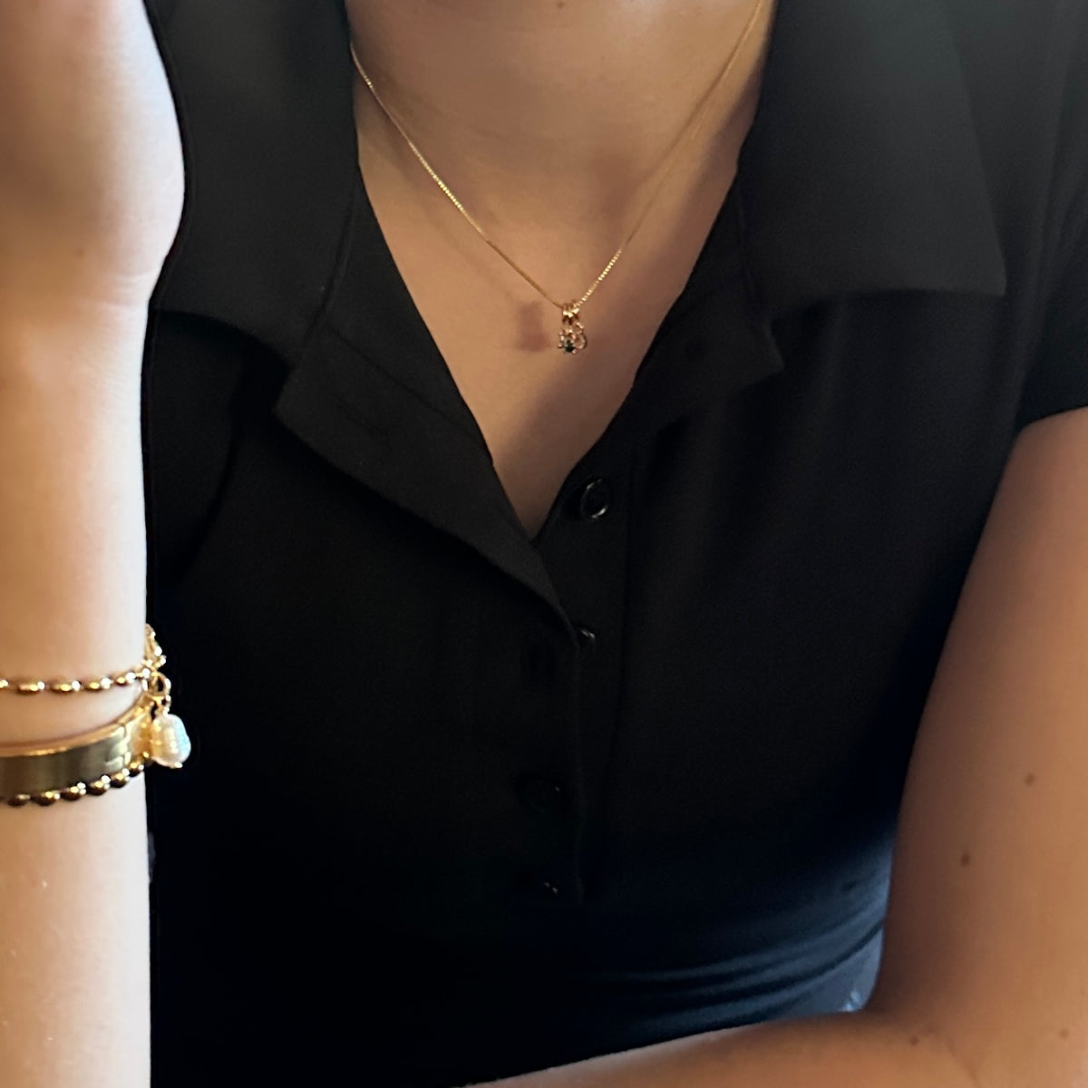 A close-up lifestyle shot of a person wearing a delicate gold necklace with a small anchor pendant, styled with a black collared top and stacked gold bracelets on the wrist.