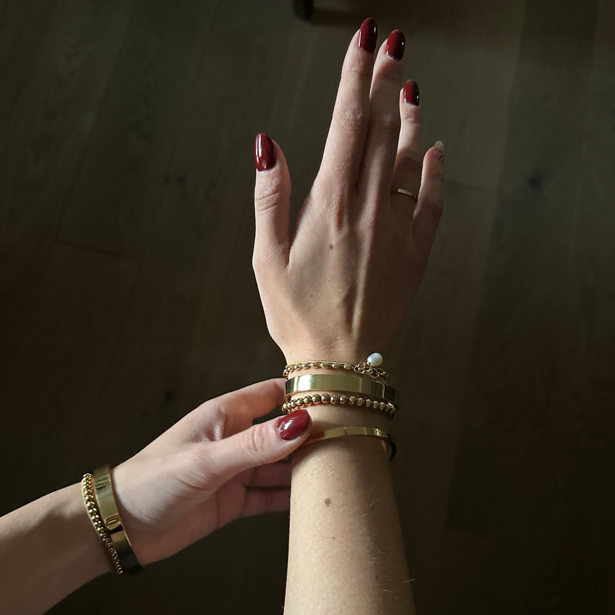A close-up of a person's hands and wrist wearing multiple gold bracelets, including a beaded bangle, a pearl accent bracelet, and a slim gold band, against a dark wooden background.
