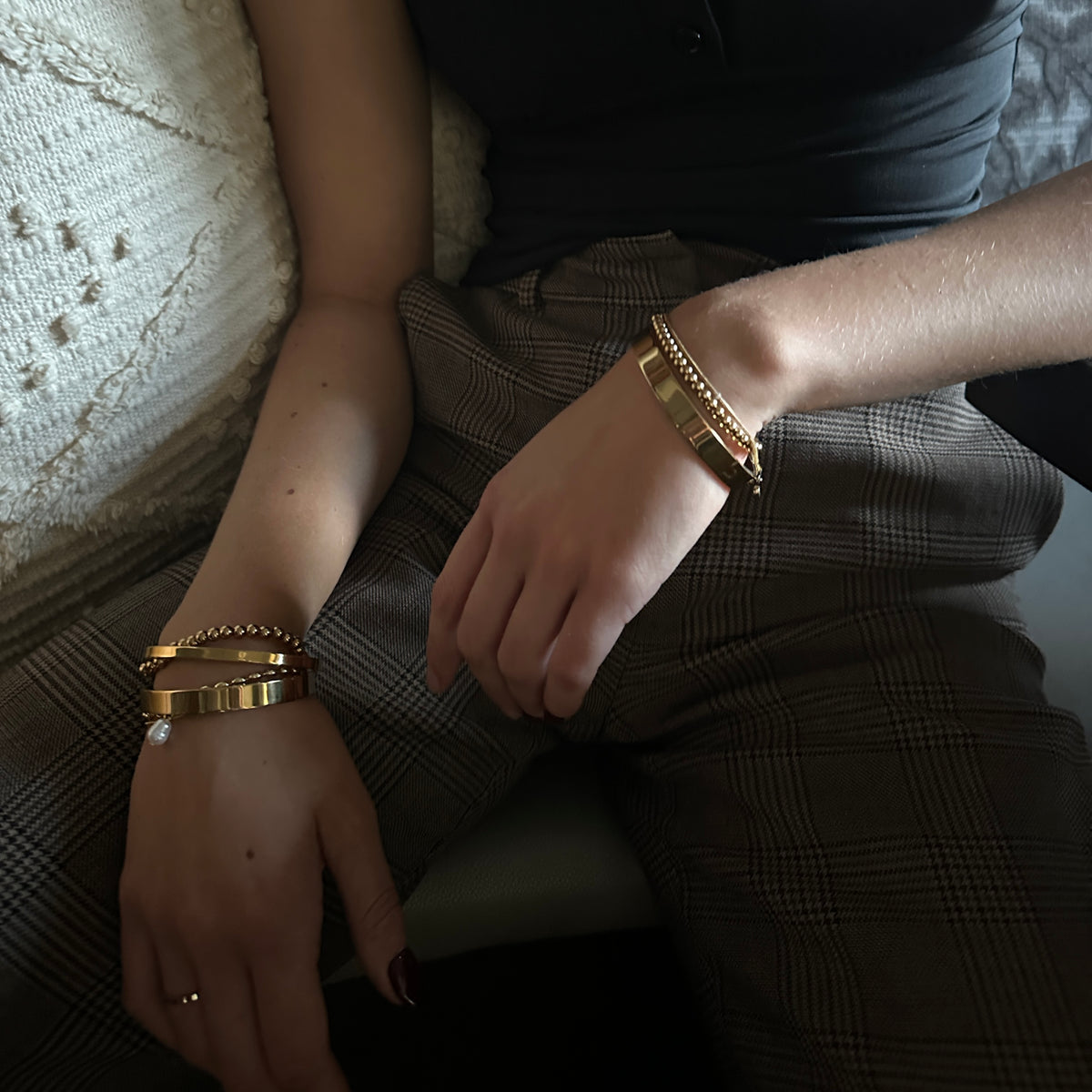 A close-up lifestyle shot of a person wearing multiple gold bracelets on both wrists, sitting on a textured cream sofa. The person is wearing a black top and brown plaid trousers.