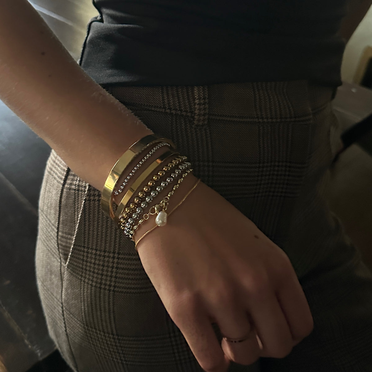 A close-up of a person's wrist wearing a stack of gold and silver bracelets, including a beaded bracelet with a small white pearl charm, paired with brown plaid trousers.