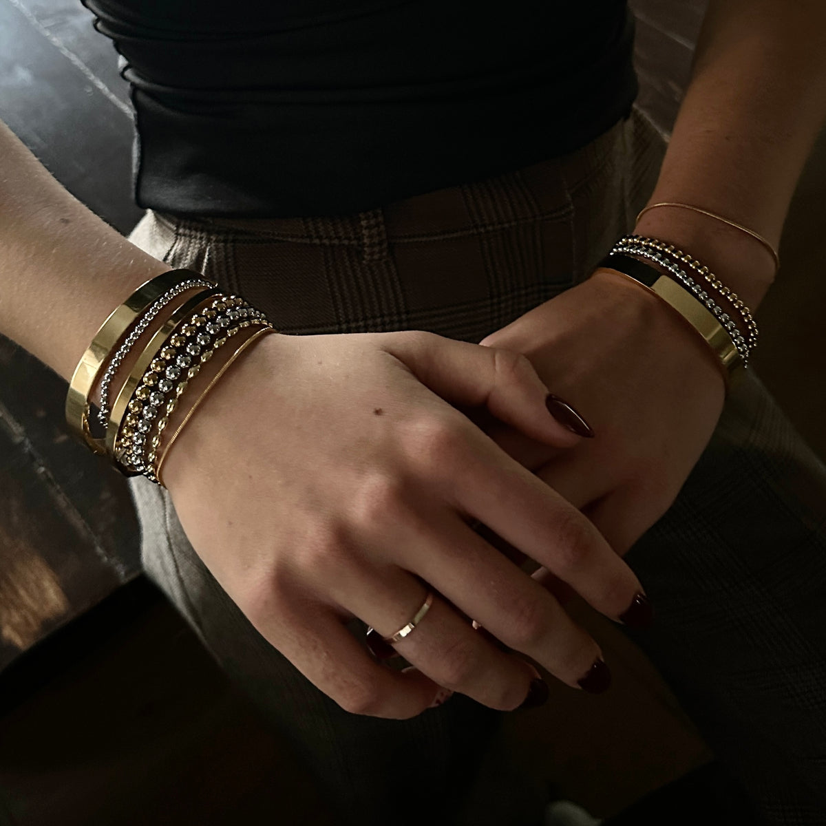 A close-up of a person's hands and arms wearing multiple stacked bracelets, including gold and silver chain styles, with a matching gold ring on the finger.