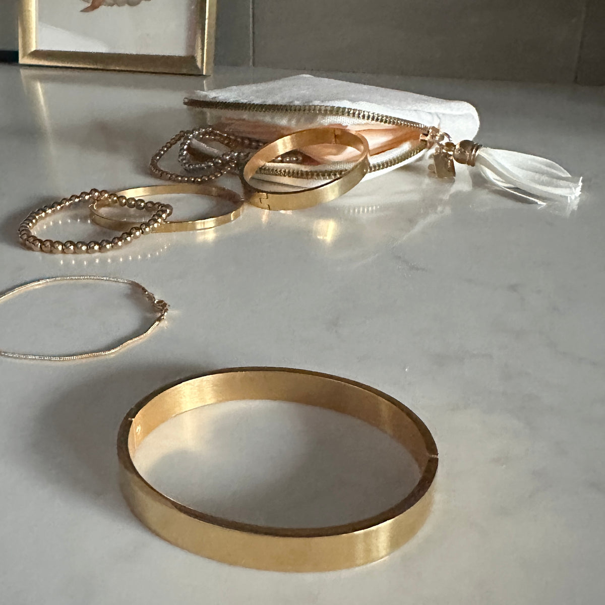 A collection of gold-toned jewelry and accessories arranged on a light marble surface, including a wide bangle, beaded bracelets, rings, a necklace, and a white zip pouch with a tassel.