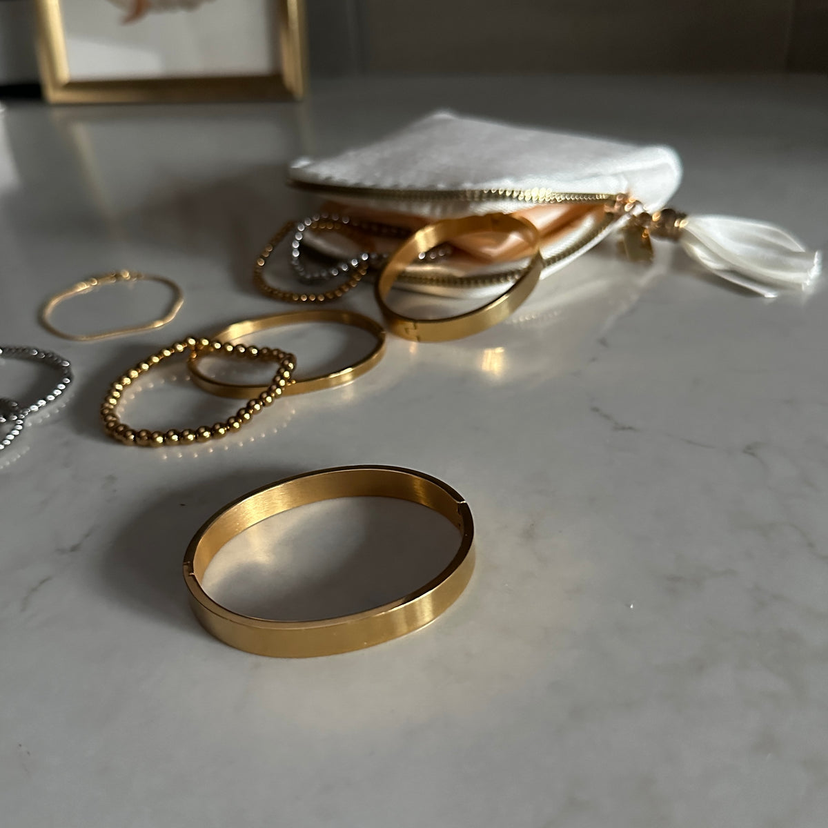 A gold bangle bracelet displayed prominently in the foreground on a light marble surface, with several other gold and silver jewelry pieces and a white pouch in the background.