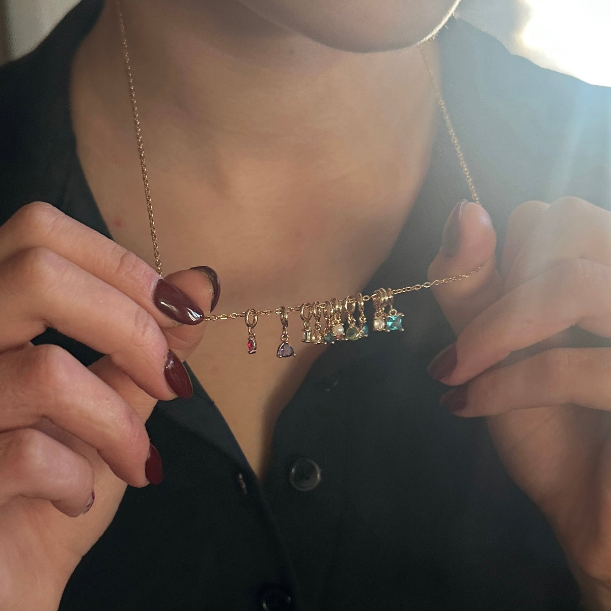 A close-up lifestyle shot of a person wearing a delicate gold chain necklace with multiple small dangling charms, including colorful gemstone drops and a small star charm. The person is wearing a black button-down shirt and holding the necklace with both hands.