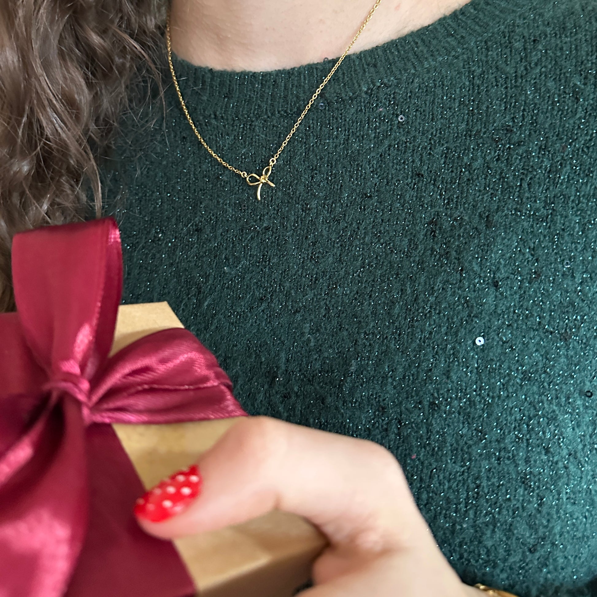 Person holding a gift box with a red ribbon, wearing a green sweater and gold necklace.