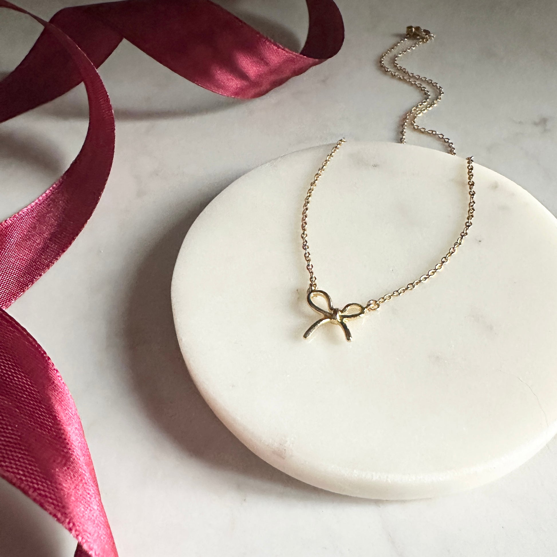 Gold necklace with a bow pendant on a marble surface with a pink ribbon.