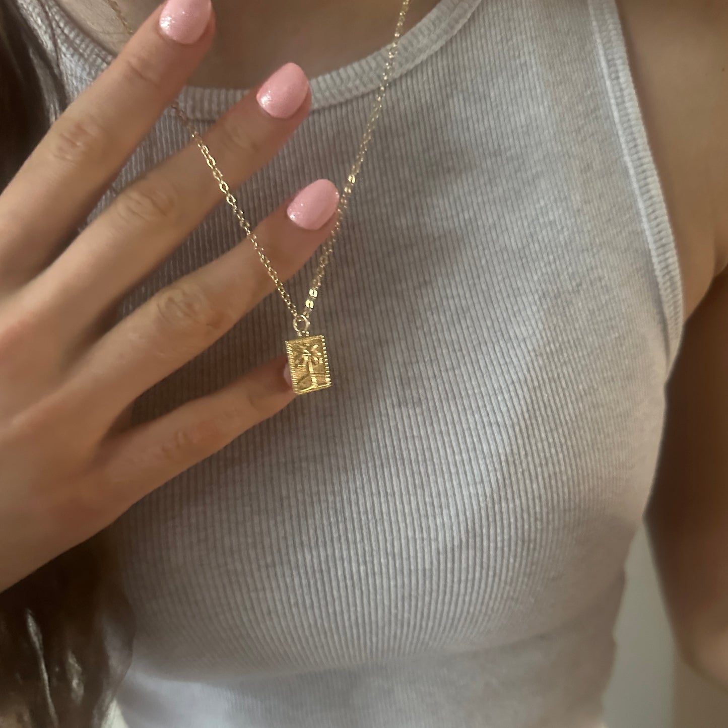 Gold necklace with a square pendant held by a hand with pink nail polish.