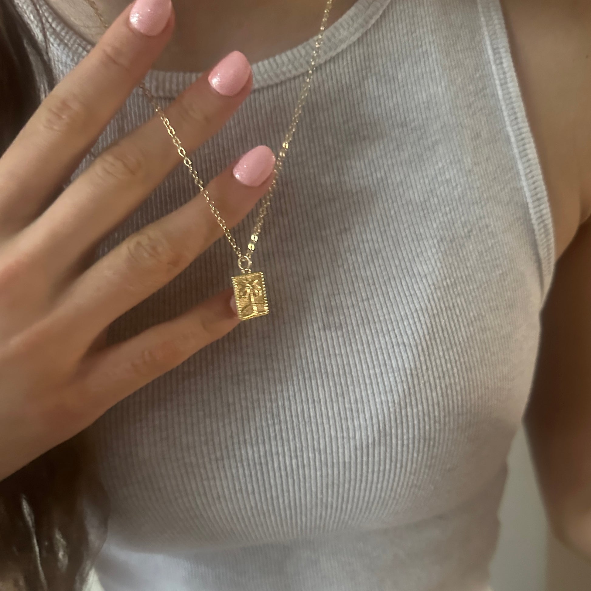 Gold necklace with a square pendant held by a hand with pink nail polish.
