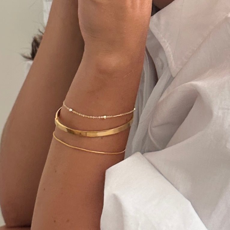 Three delicate gold bracelets stacked on a person's wrist, including a thin beaded chain, a smooth bangle, and a thin wire bracelet, paired with a white shirt.