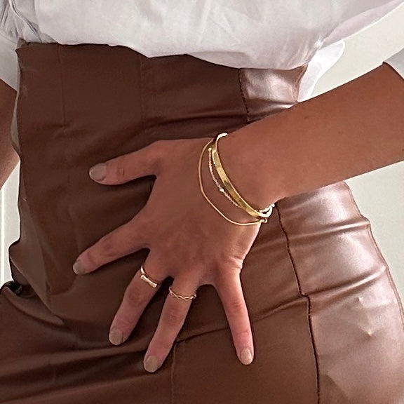 A close-up of a person's hand wearing delicate gold rings and stacked gold bracelets, resting on a brown faux-leather surface.