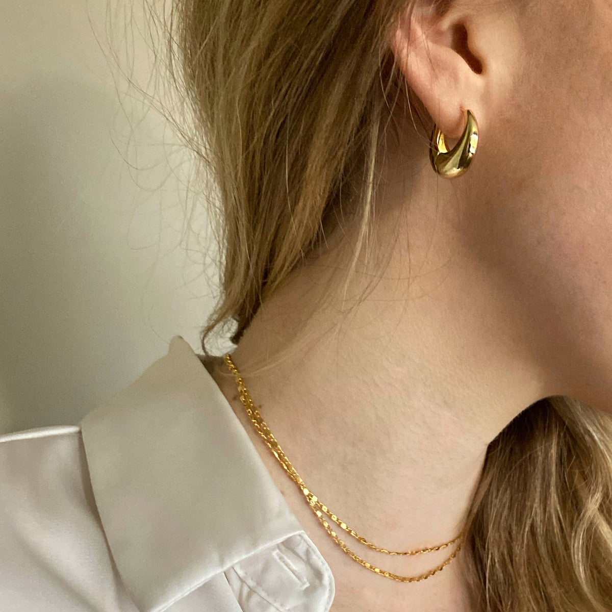 A close-up lifestyle shot of a model wearing a polished gold crescent-shaped hoop earring and a delicate gold chain necklace, paired with a white collared shirt.